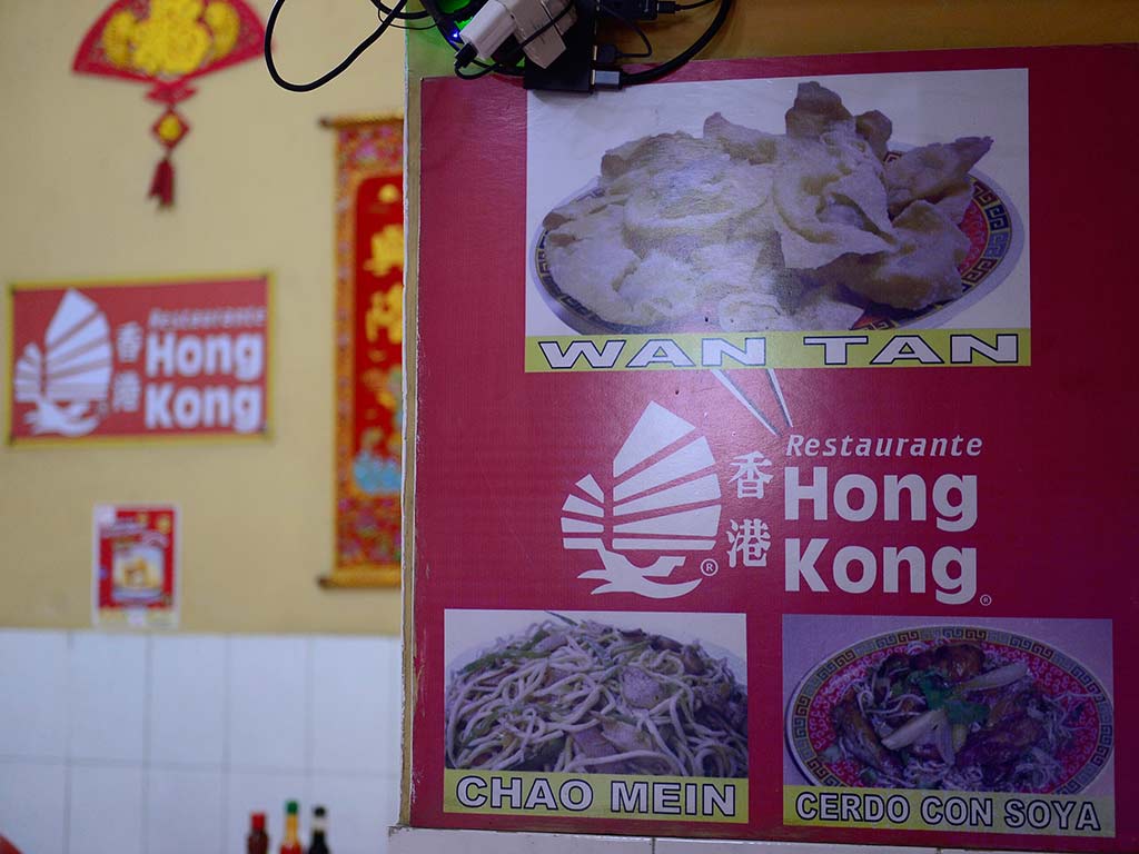 Vista de cerca de varios menús impresos y coloridos en la pared de un restaurante. Tienen fotos de platos chinos como "ARROZ CANTONES", "SOPA LOMITO", "1/2 COMBINADO HAWAII", y "SOPA WANTAN". Los menús tienen texto en español y chino, y un logotipo con el nombre "Restaurante Hong Kong".