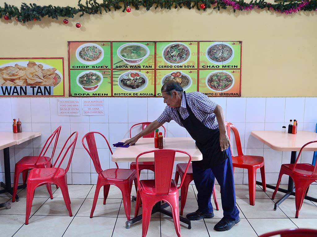 Un hombre mayor con una camisa de cuadros y un delantal azul está agachado, limpiando una mesa blanca con un paño azul en un restaurante chino. A su alrededor hay sillas de metal rojas. En la pared del fondo, hay menús visuales grandes con fotos de platos como "CHOP SUEY", "SOPA WAN TAN" y "CHAO MEIN". Una guirnalda verde decora la parte superior de la pared.