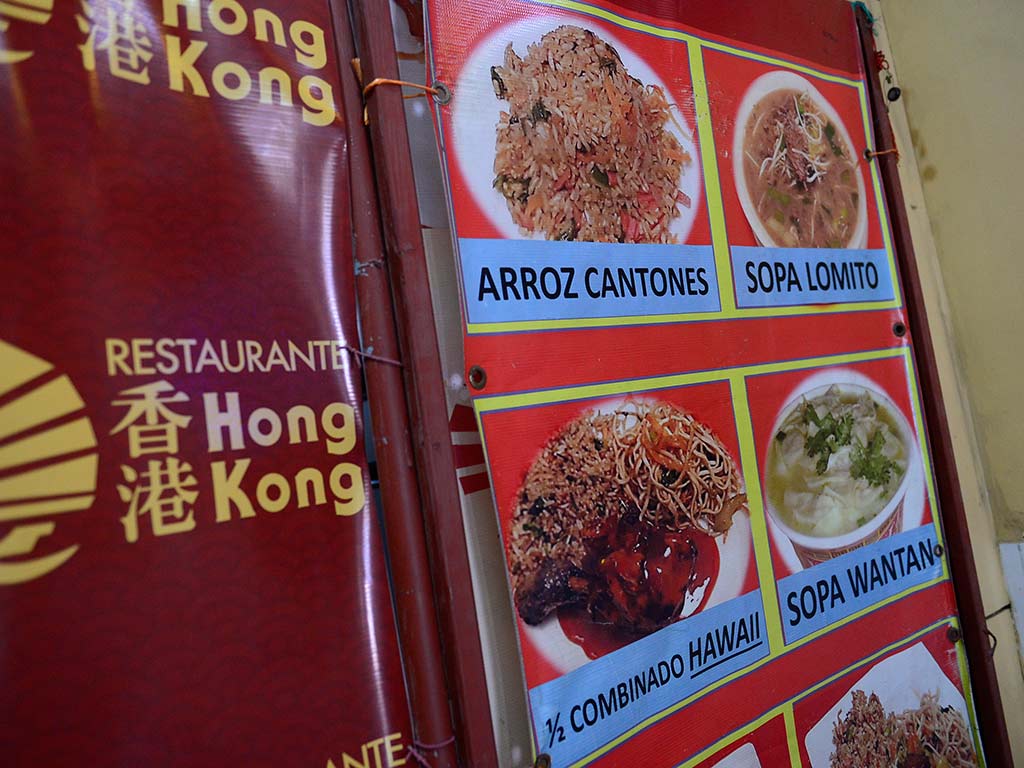 Una pared de restaurante cubierta con un gran menú impreso y colorido. Muestra fotos y nombres de platos como "CHULETA DE TERNERA", "SOPA WAN TAN", "WAN TAN", "SOPA MEIN", "POLLO CON SEMILLAS", "CAMARONES EMPANIZADOS", "BISTEK DE AGUJA" y "1/4 DE POLLO". En la parte inferior derecha, hay un logotipo de "Restaurante Hong Kong" y texto celebrando "50 años de Tradición". El fondo es de baldosas blancas.
