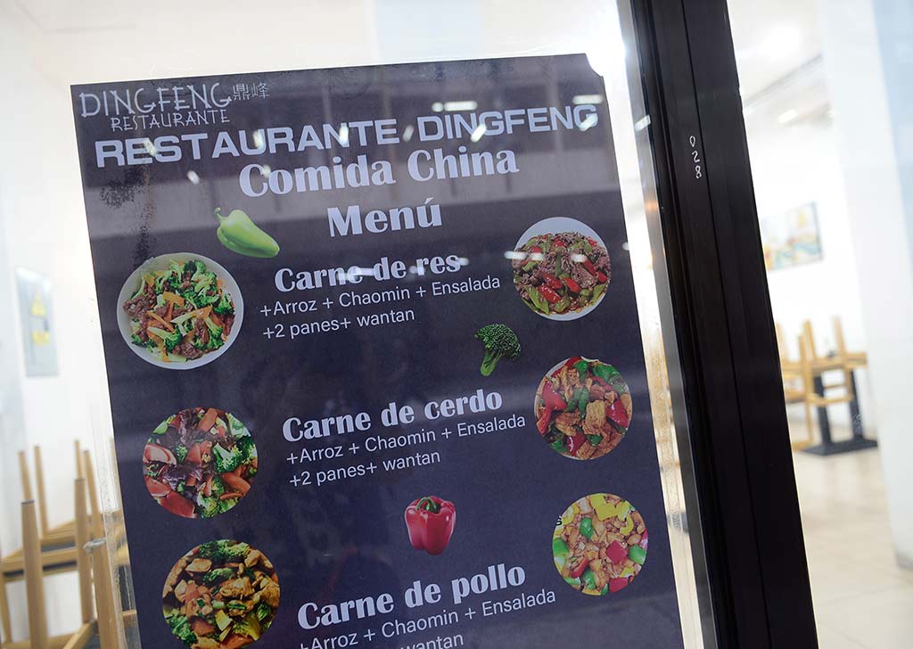 Primer plano de un menú impreso y colorido pegado en la vidriera de cristal de un restaurante. El menú tiene el logotipo "DINGFENG RESTAURANTE" en la parte superior y el título "Restaurante Ding Feng Comida China Menú". A continuación, presenta tres combos ilustrados con fotos apetitosas de platos chinos: "Carne de res", "Carne de cerdo" y "Carne de pollo", cada uno con texto que detalla los acompañamientos como arroz, chao mein, ensalada, pan y wantan. El menú está decorado con imágenes de pimientos y brócoli. A la derecha, un reflejo muestra sillas de madera y una mesa en el interior del restaurante.