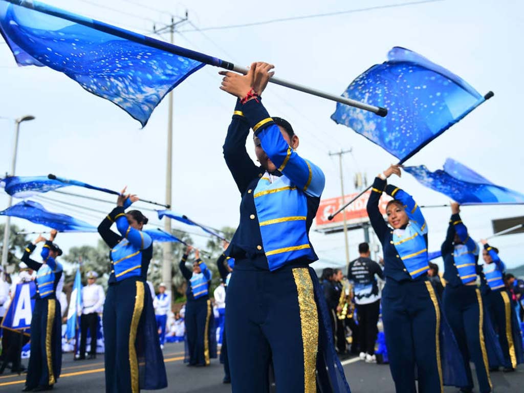 Grupo de estudiantes vestidas con uniforme azul y dorado ondeando banderas celestes en una calle durante un desfile.