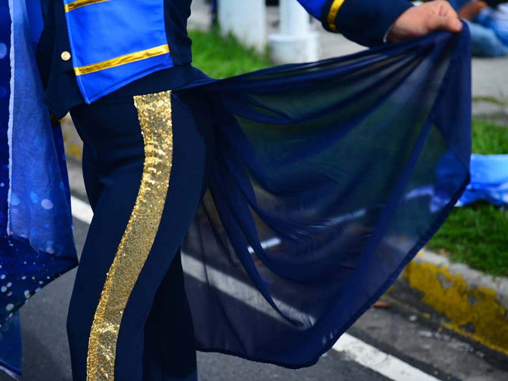Fotografía en primer plano de la pierna y el costado de una banderista del Liceo Cristiano, mostrando la franja dorada vertical en el pantalón de vestir azul oscuro y la capa traslúcida que se extiende por detrás. El borde de la bandera azul con destellos es visible a la izquierda.