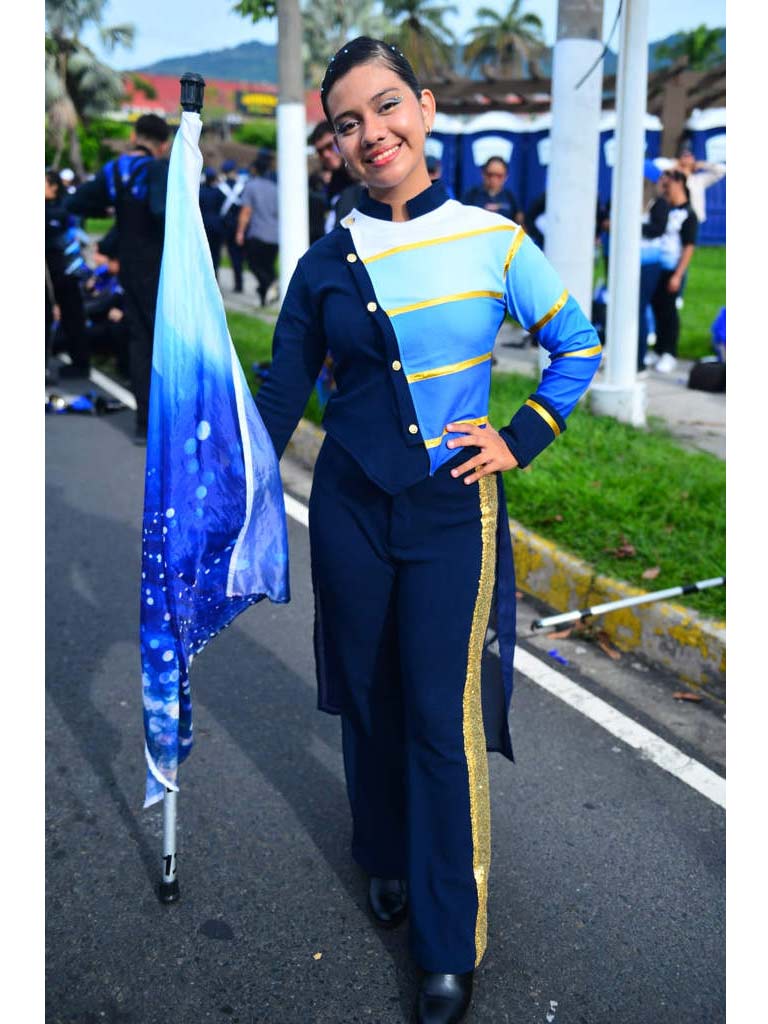 Fotografía de cuerpo entero de una joven banderista sonriente del Liceo Cristiano Reverendo Juan Bueno Central. Viste un uniforme sastre en tonos azul, blanco y celeste con detalles dorados. Sostiene una bandera azul celeste con una mano y tiene la otra en la cintura. El fondo muestra una calle pavimentada y otros estudiantes desenfocados.