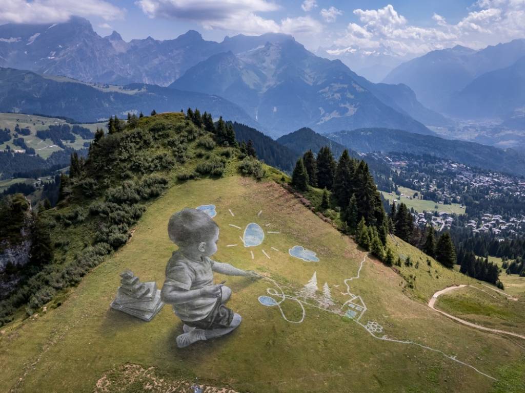 Perspectiva aérea de un mural gigante sobre la hierba con un niño pintando un sol, árboles y una casa en la montaña.