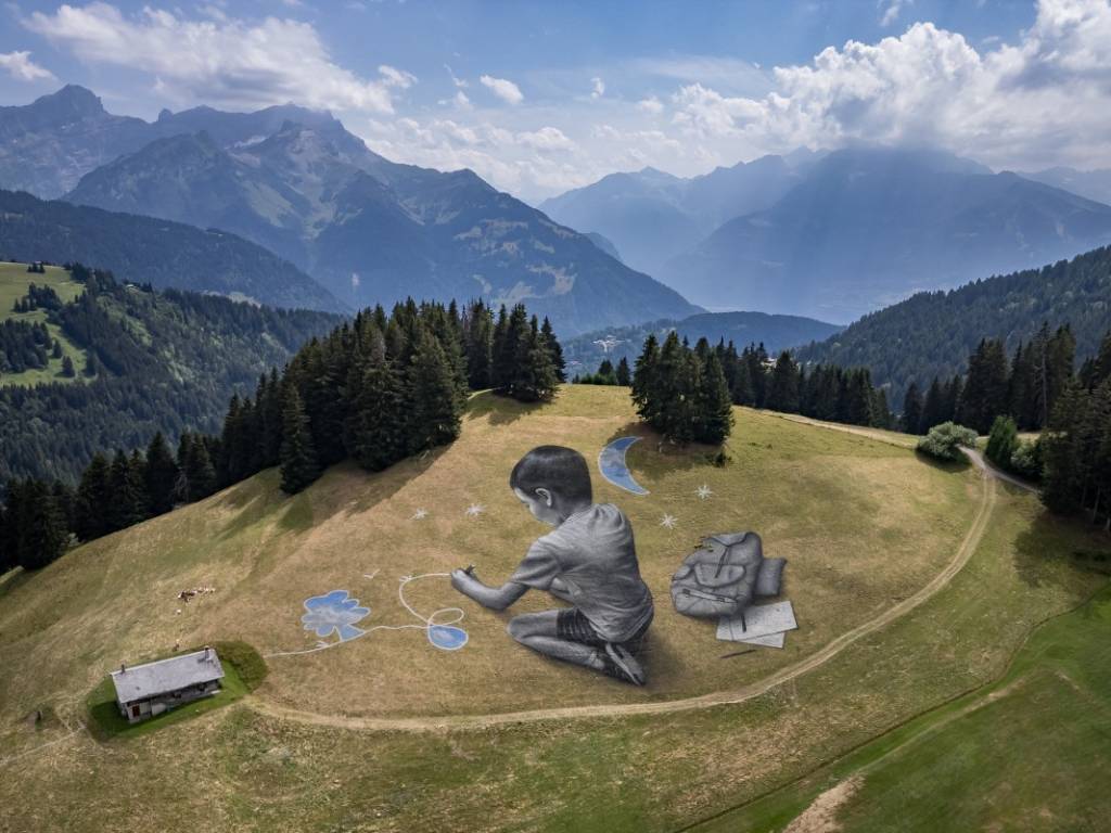 Vista panorámica de un niño gigante pintado en una ladera verde, dibujando estrellas y la luna sobre el césped.
