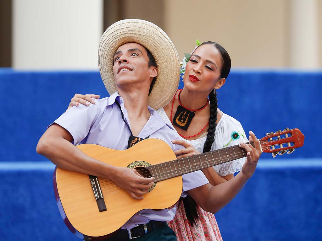 Un hombre con sombrero de paja y camisa lila toca una guitarra acústica mientras una mujer detrás de él, con un vestido folclórico blanco y rojo y flores en el cabello, sonríe y lo abraza.