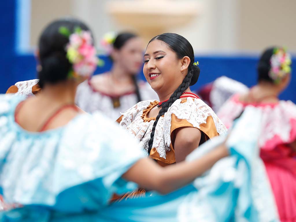 Una joven con flores rosas en el cabello y un vestido tradicional blanco y rojo sonríe mientras baila, de pie frente a otra bailarina de espaldas que lleva un vestido azul claro.