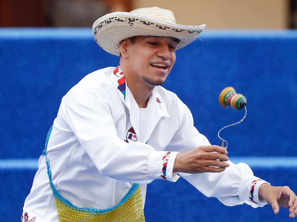 Un joven con sombrero de paja y camisa blanca tradicional sonríe mientras manipula un capirucho de madera con una cuerda.