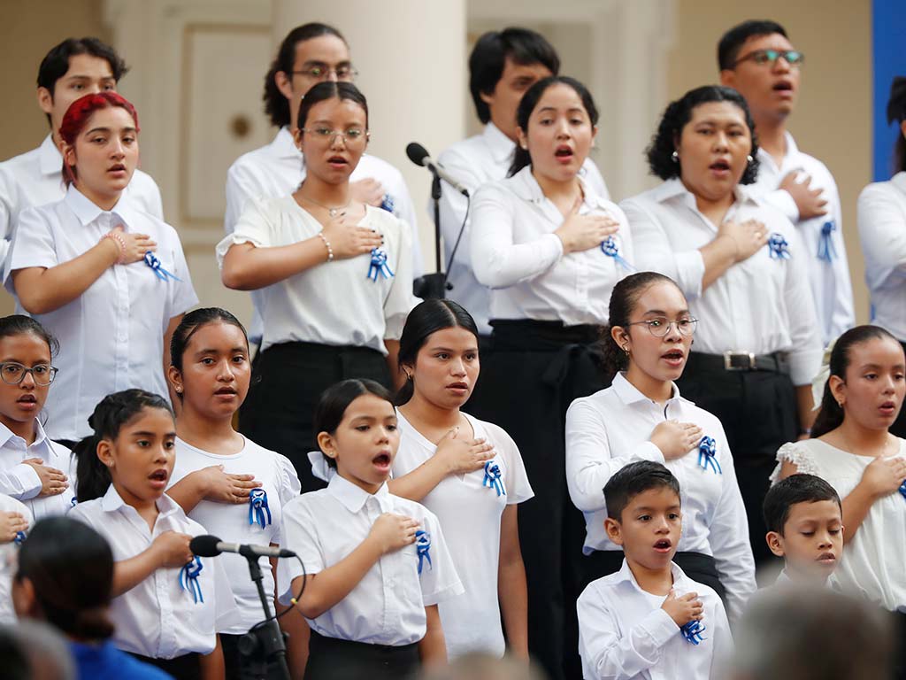 Un grupo de estudiantes de coro, vestidos con camisas blancas y escarapelas azul y blanco, cantan juntos con la mano derecha sobre el corazón.