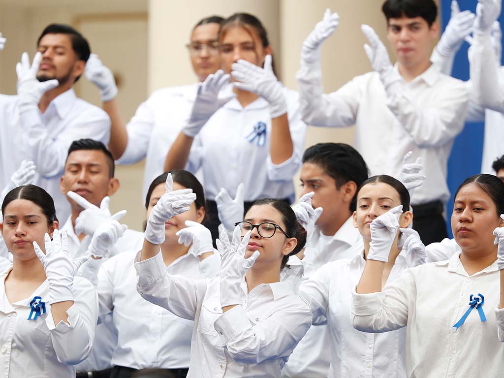 Un grupo de estudiantes vestidos de blanco, con guantes blancos, realizan gestos con las manos al unísono, como parte de una presentación en lenguaje de señas.