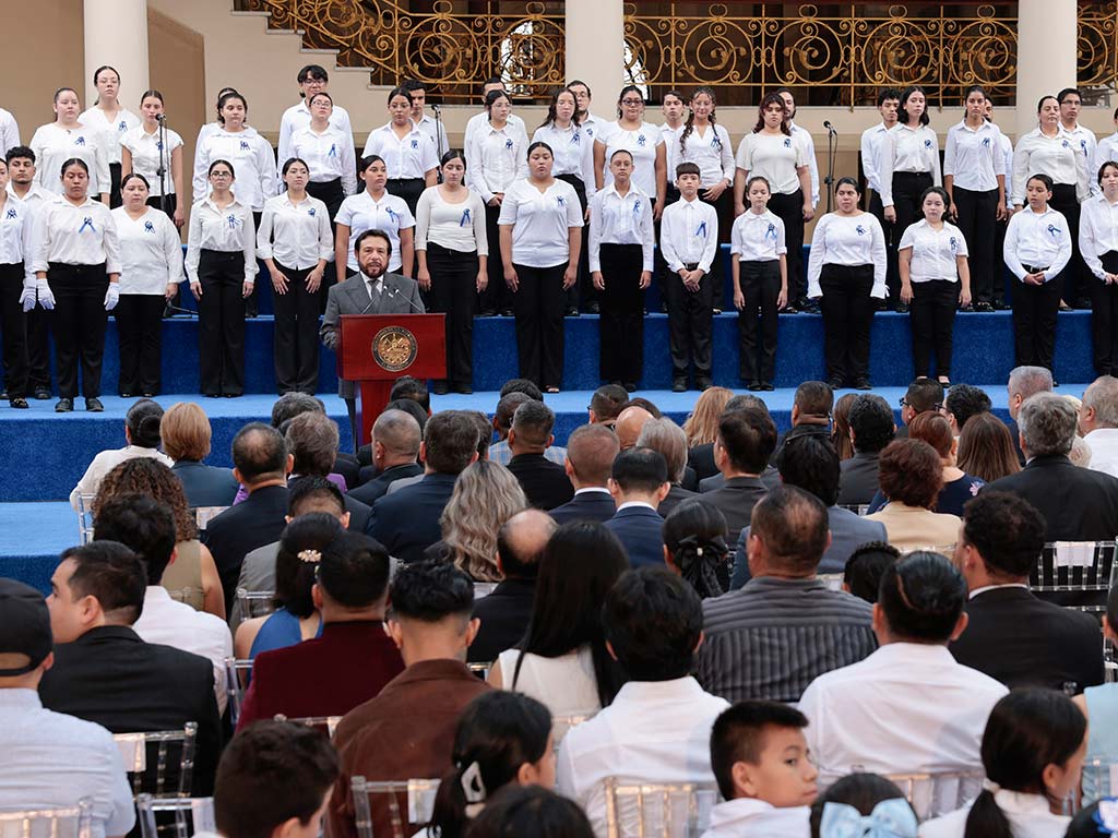 Primer plano de Félix Ulloa, Vicepresidente de El Salvador, con un traje gris, hablando en un podio con micrófono. Detrás de él, estudiantes del coro.