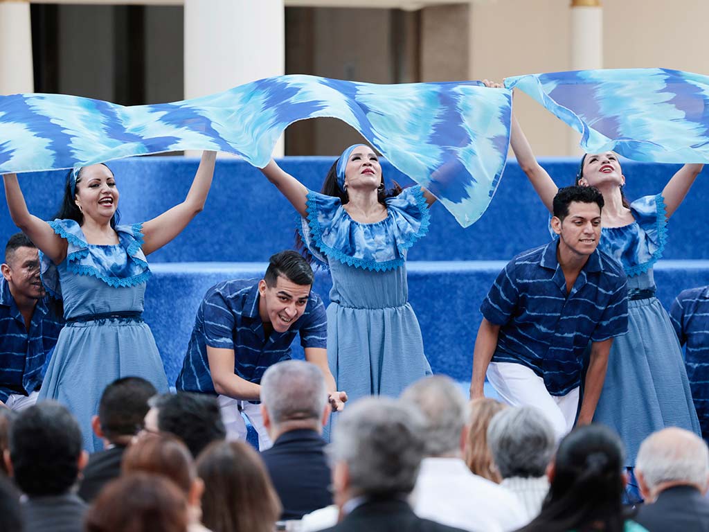 Un grupo de bailarines vestidos con trajes azul y blanco ondean grandes telas de tela azul claro y blanco por encima de sus cabezas, frente a la audiencia.