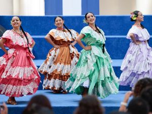 Cuatro mujeres con vestidos folclóricos tradicionales de colores rosa, naranja, verde y lila posan juntas con las manos en las caderas, sonriendo.