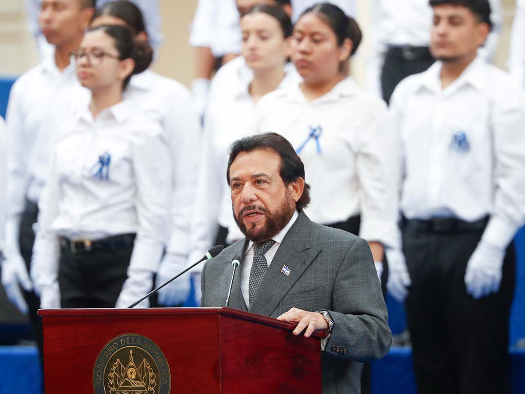 Félix Ulloa, Vicepresidente de El Salvador, pronuncia un discurso en un podio de madera con el escudo de El Salvador, frente a una gran audiencia y el coro estudiantil detrás de él.