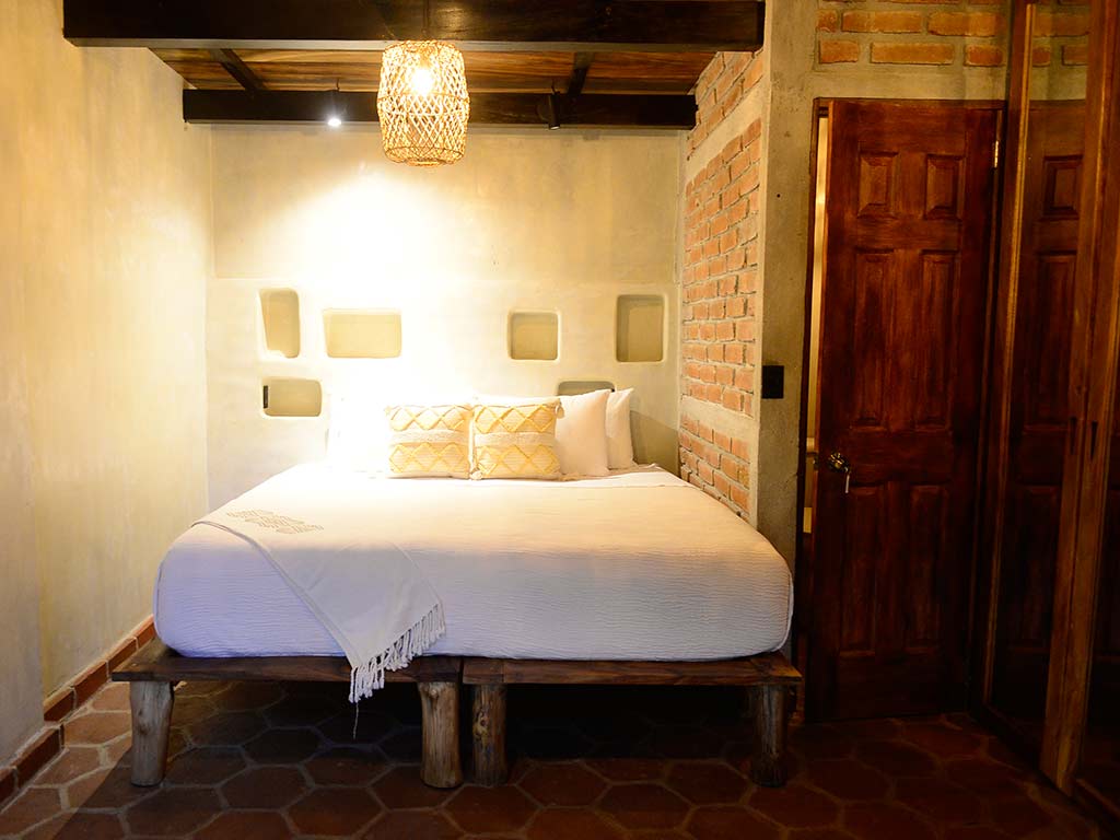 Dormitorio acogedor en Casona Tunan con cama king, paredes de ladrillo visto, detalles en barro y lámparas artesanales tejidas.