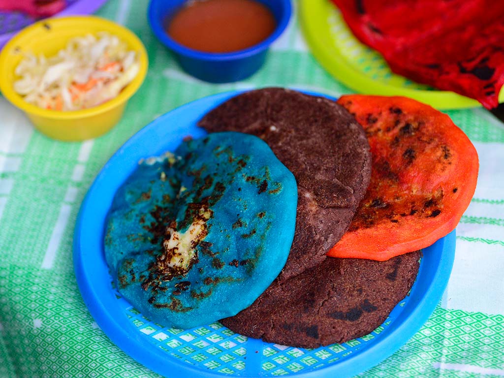 Plato con pupusas salvadoreñas variadas incluyendo la pupusa negrita hecha con masa de frijol refrito y pupusas azul y naranja.