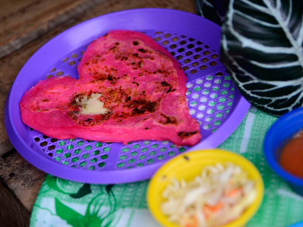 Pupusa artesanal rosada en forma de corazón con quesillo derretido, acompañada de curtido y salsa tradicional salvadoreña.