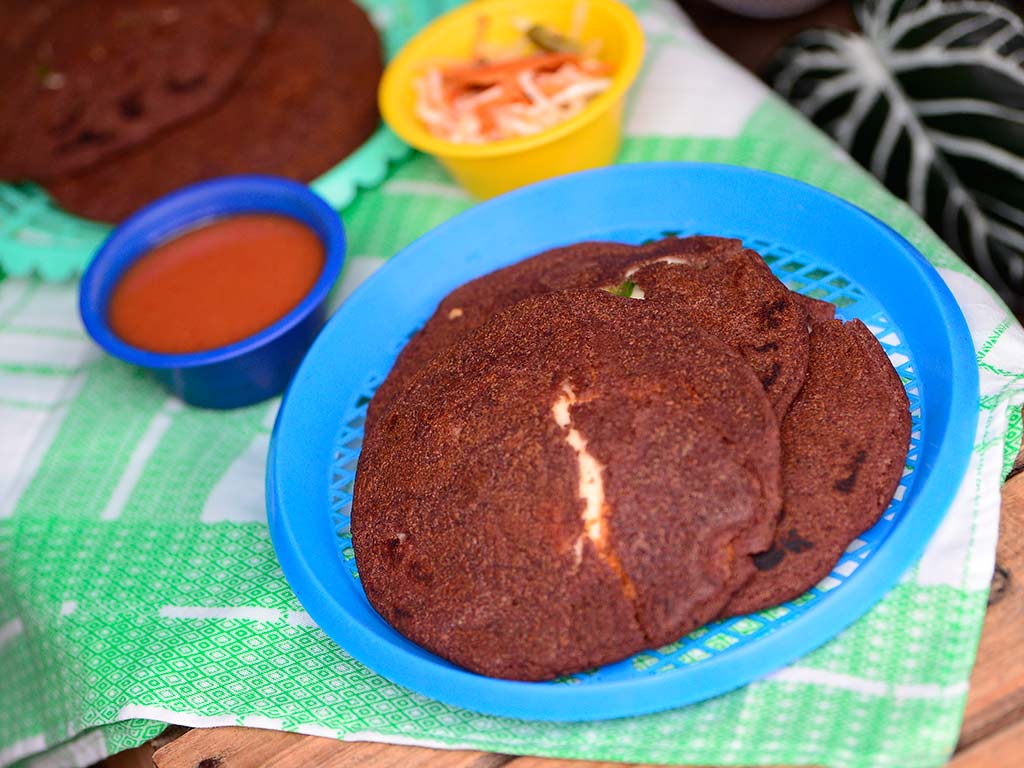 Plato de pupusas negritas de masa de frijol acompañadas con salsa de tomate y curtido salvadoreño.