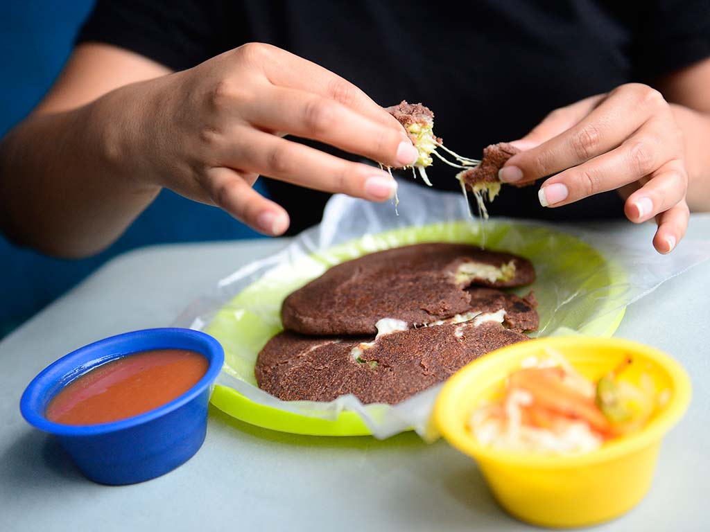 Comensal abriendo una pupusa de masa de frijol refrito derramando queso derretido en su interior.
