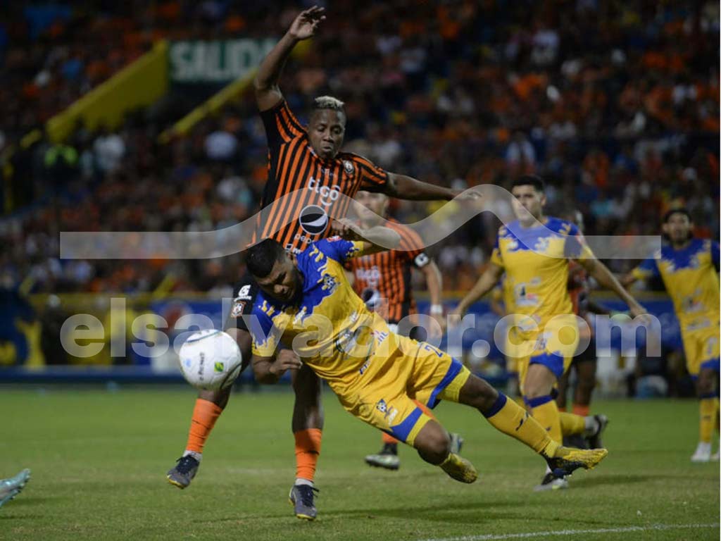 Una fotografía a color que muestra a varios jugadores de Jocoro FC abrazándose y celebrando un gol en un estadio con el cielo nublado de fondo. Visten uniformes amarillos y negros.