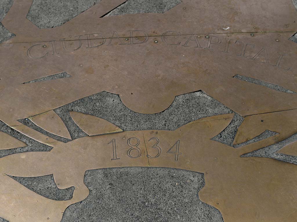 Detalle del escudo metálico en el suelo que conmemora el año 1834, fecha clave en la que San Salvador se consolidó como capital de la República Federal de Centroamérica.