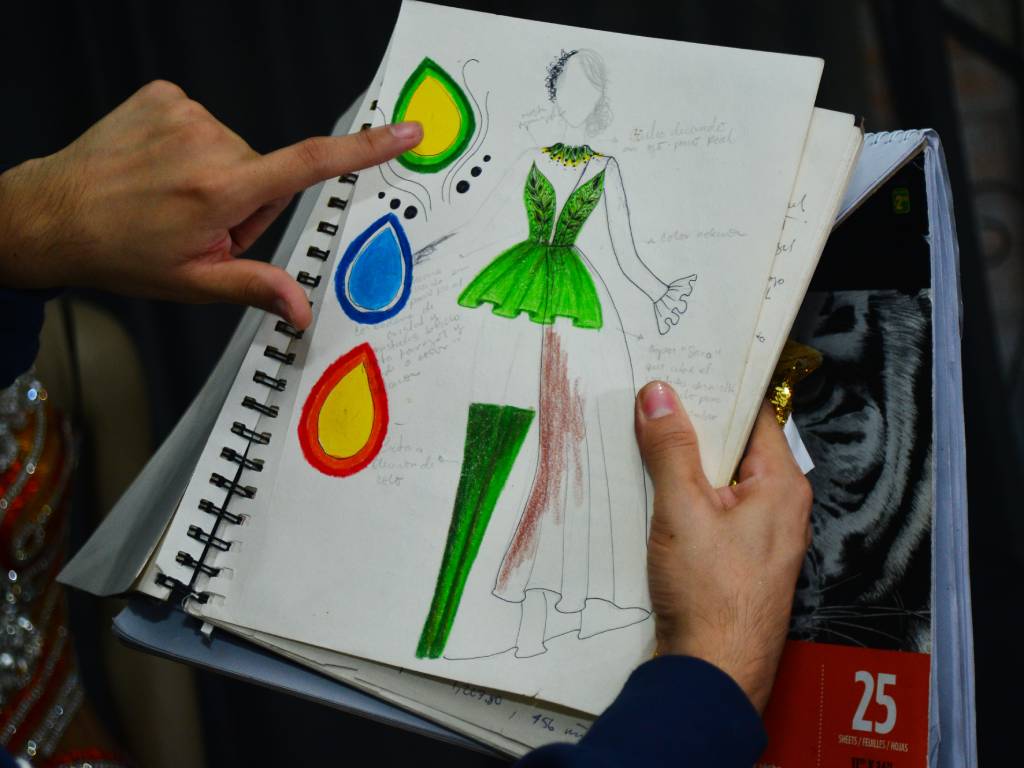 Roni Matías sosteniendo y mostrando varias hojas con bocetos ilustrados de trajes de cachiporra dibujados a mano.