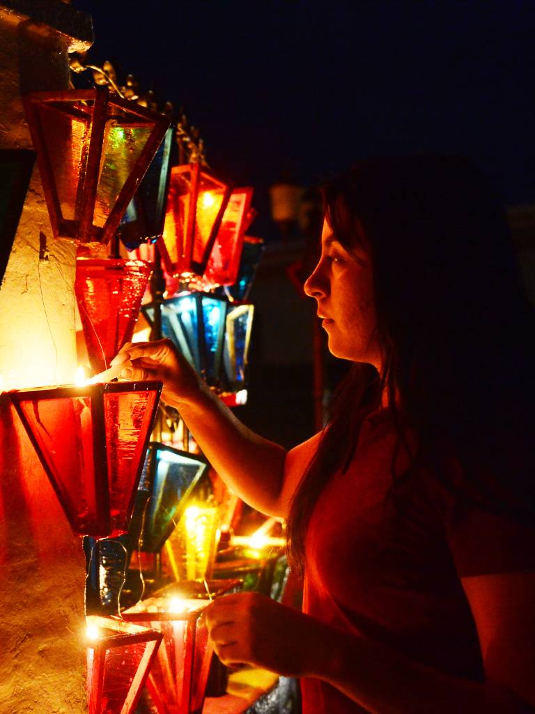 Personas encendiendo velas dentro de farolitos colocados en fachadas y árboles al atardecer.