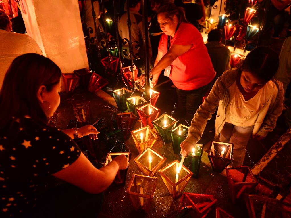 Una mujer con camisa roja, otra con blusa de estrellas y una niña pequeña inclinadas encendiendo velas en farolitos de celofán verde, rojo y amarillo colocados sobre el suelo.