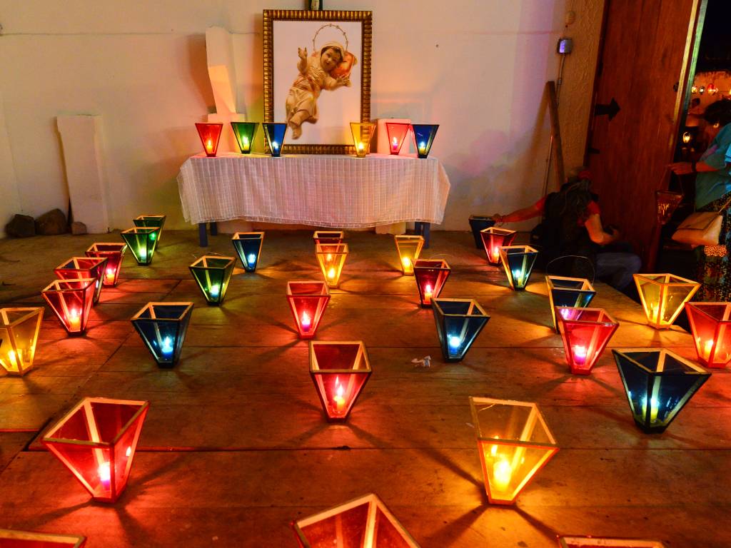 Cuadro de la Virgen Niña sobre una mesa vestida con mantel blanco, rodeado en el piso por farolitos encendidos de colores rojo, azul y amarillo.