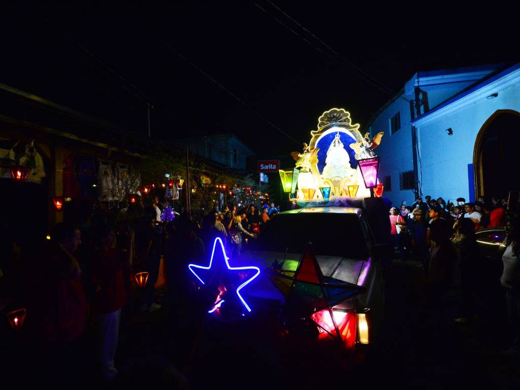 Procesión festiva con la pequeña imagen de la Virgen Niña avanzando por calles iluminadas por farolitos y acompañada por feligreses.