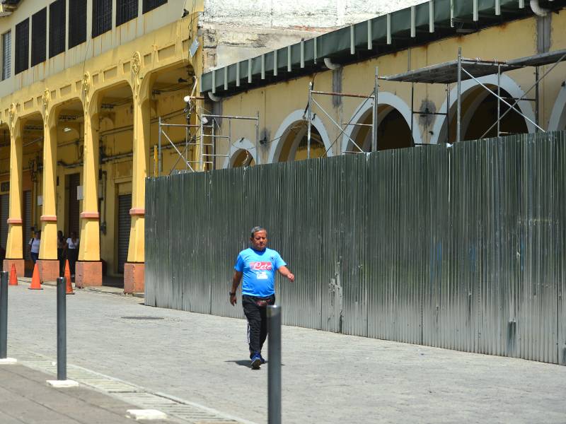 Vista que muestra el Portal Sagrera cubierto por láminas metálicas a la derecha, en ángulo hacia el Portal La Dalia a la izquierda, que tiene una fachada amarilla y roja diferente, formando un corredor continuo.