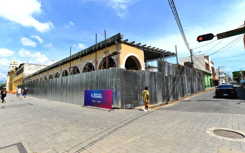 Vista de esquina del Portal Sagrera cubierto por láminas metálicas de construcción plateadas y andamios, bajo un cielo azul con nubes en San Salvador.