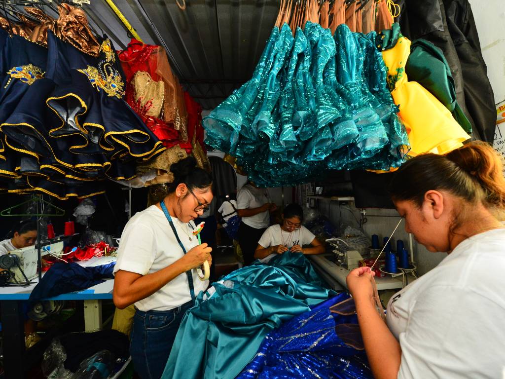 Mujeres trabajando en un taller de costura: unas operan máquinas industriales y otras realizan bordados manuales en trajes azules.