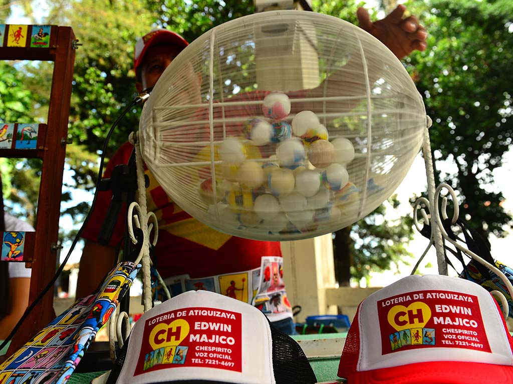 Primer plano de una tómbola esférica metálica repleta de pelotas de plástico blancas y gorras promocionales al frente.
