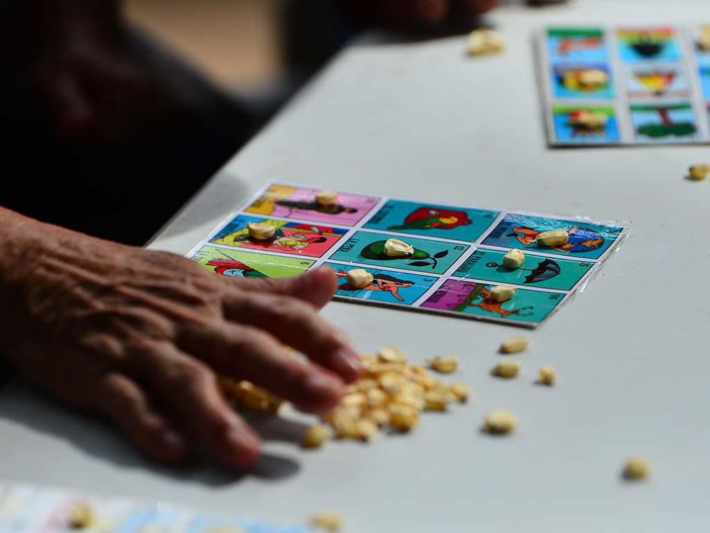 Primer plano de una mano colocando granos de maíz sobre las casillas de un cartón colorido de lotería.