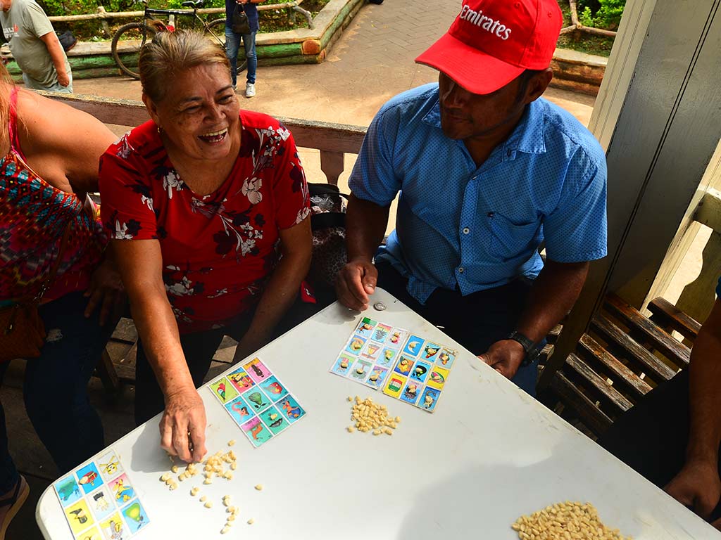 Mujer sonriendo alegremente mientras juega a la lotería sentada junto a otro participante en una mesa.