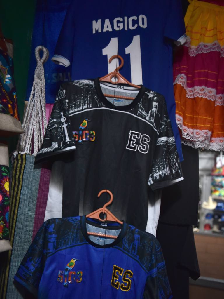 Camisetas negras y azules exhibidas con diseños alusivos a El Salvador y al Mágico González.