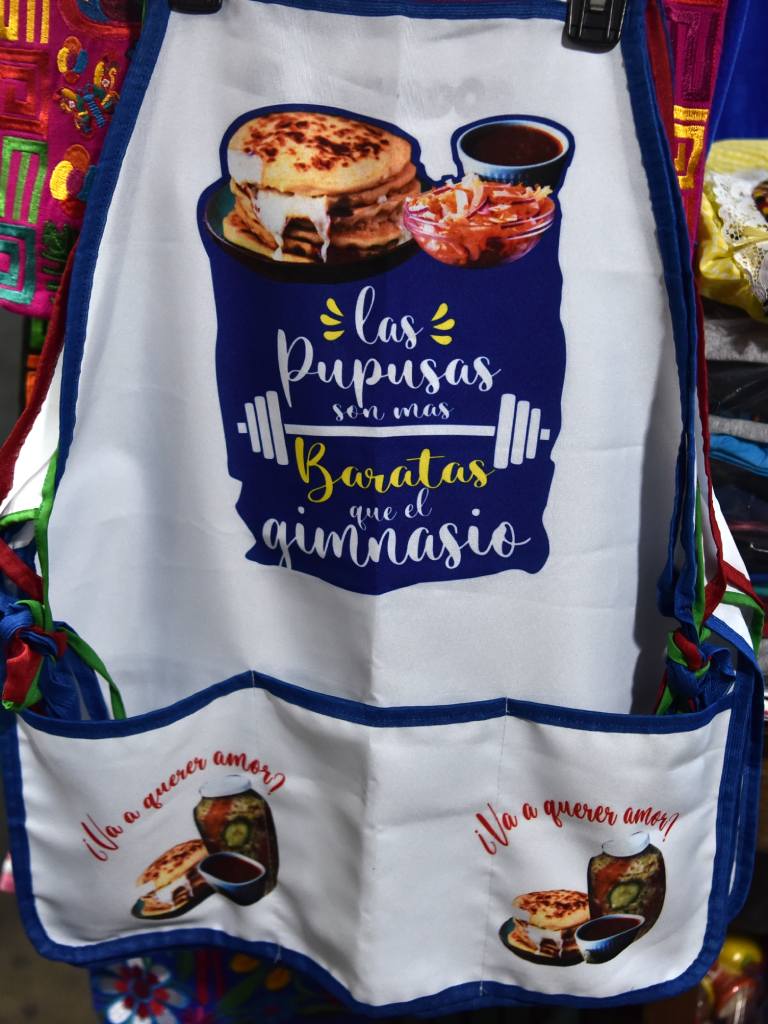 Mandil decorado con un plato de pupusas y frases humorísticas sobre la gastronomía local.