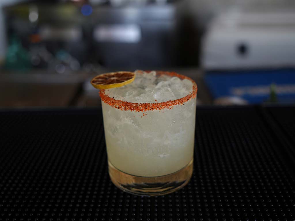 Cóctel artesanal con hielo, rodaja de limón deshidratado y borde decorado con chile en polvo en NAWI Beach House.