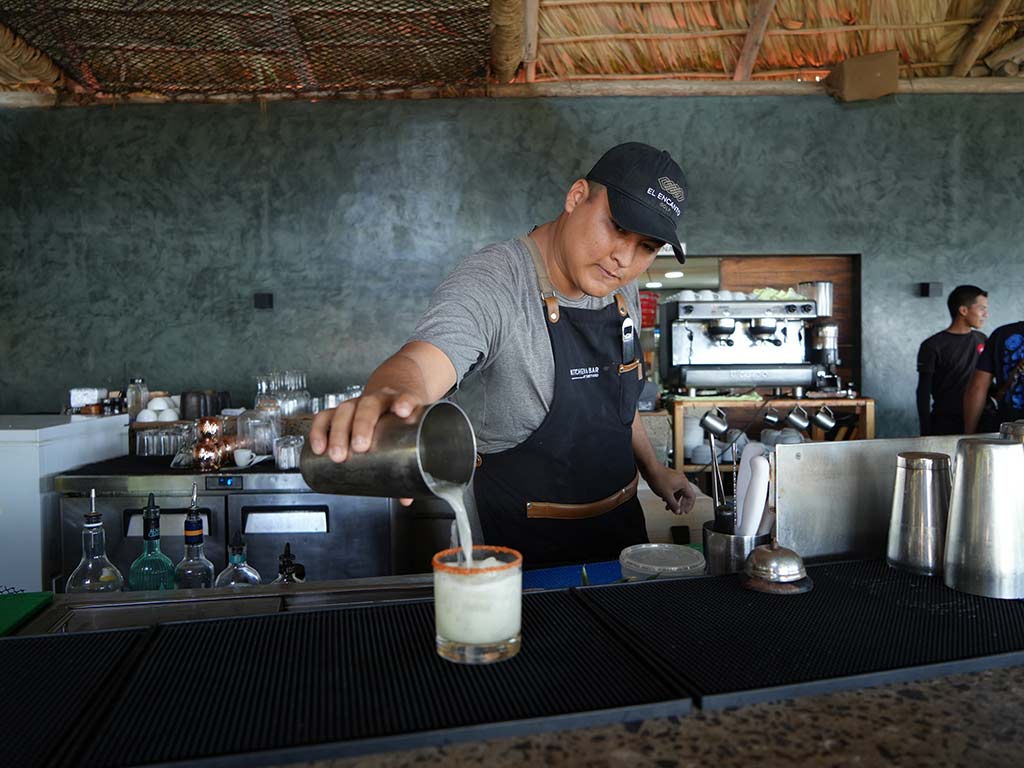 Bartender sirviendo un cóctel artesanal en vaso con borde escarchado en la barra tropical de NAWI Beach House Mizata.