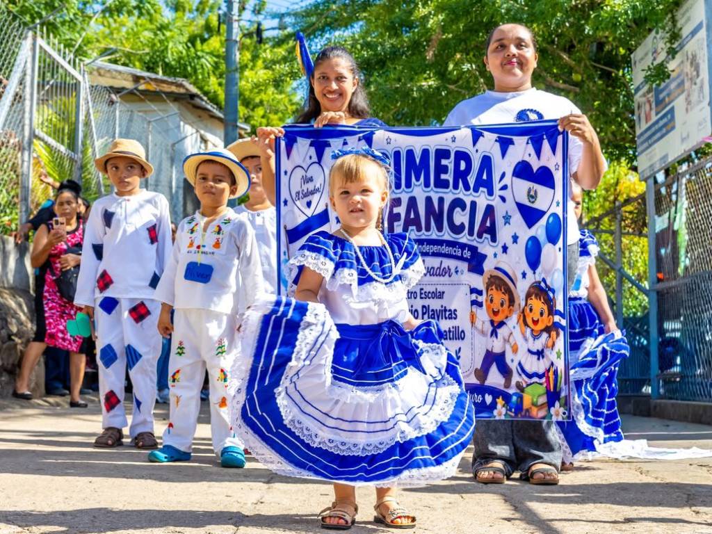 Maestros y padres de familia caminan al lado de niños pequeños mientras marchan ordenadamente por la calle.