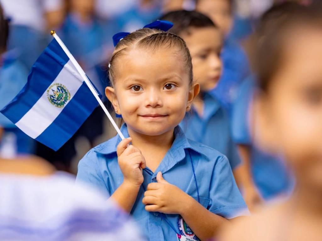 Niña vestida con uniforme escolar sonríe hacia la cámara mientras sostiene una pequeña bandera nacional de El Salvador.