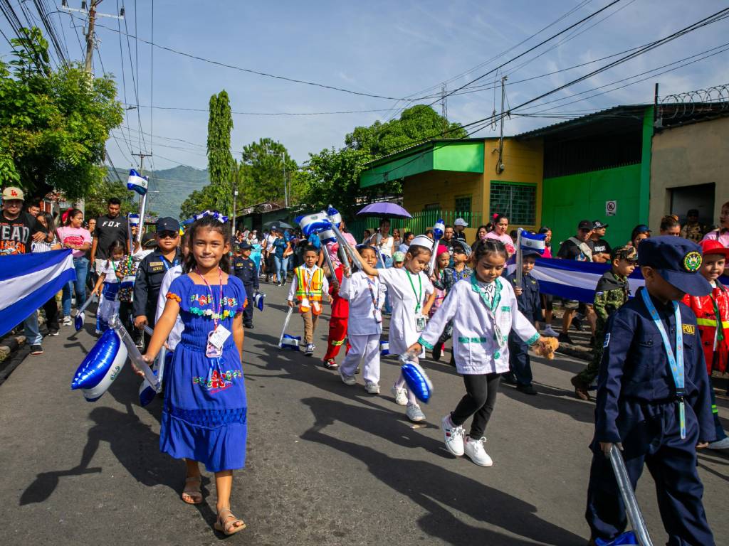 Niños y niñas luciendo trajes de doctor, bombero y otras profesiones marchan ordenadamente en un desfile sobre la calle.