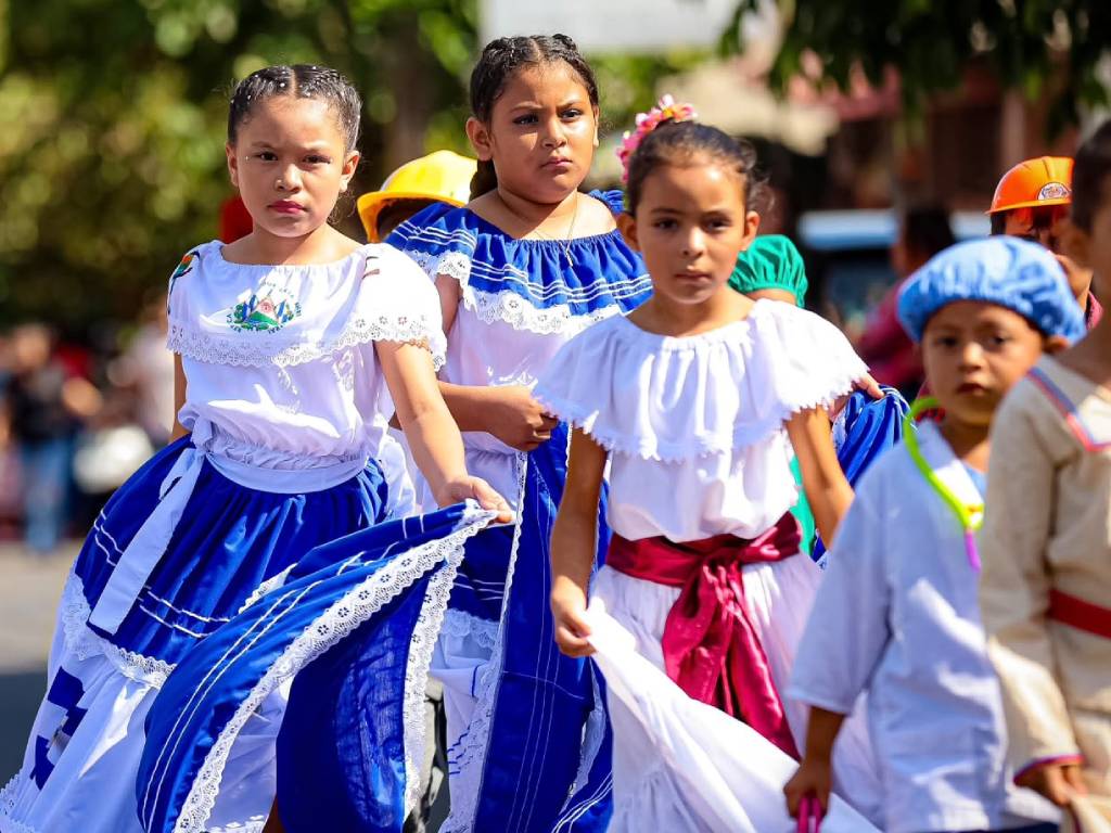 Estudiantes luciendo vestidos folclóricos tradicionales marchan en fila sobre la calle durante una presentación cultural.