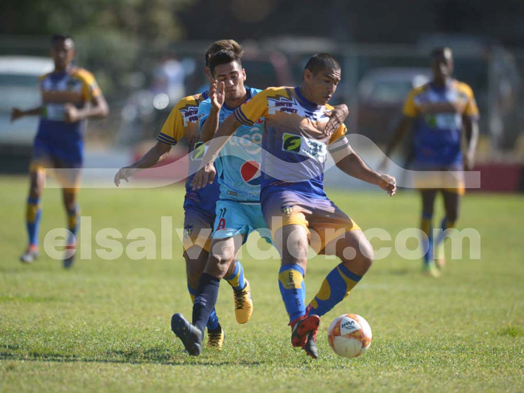 Una foto a color de un jugador de Pasaquina FC con su uniforme azul y amarillo, corriendo con el balón en un estadio soleado con montañas de fondo.