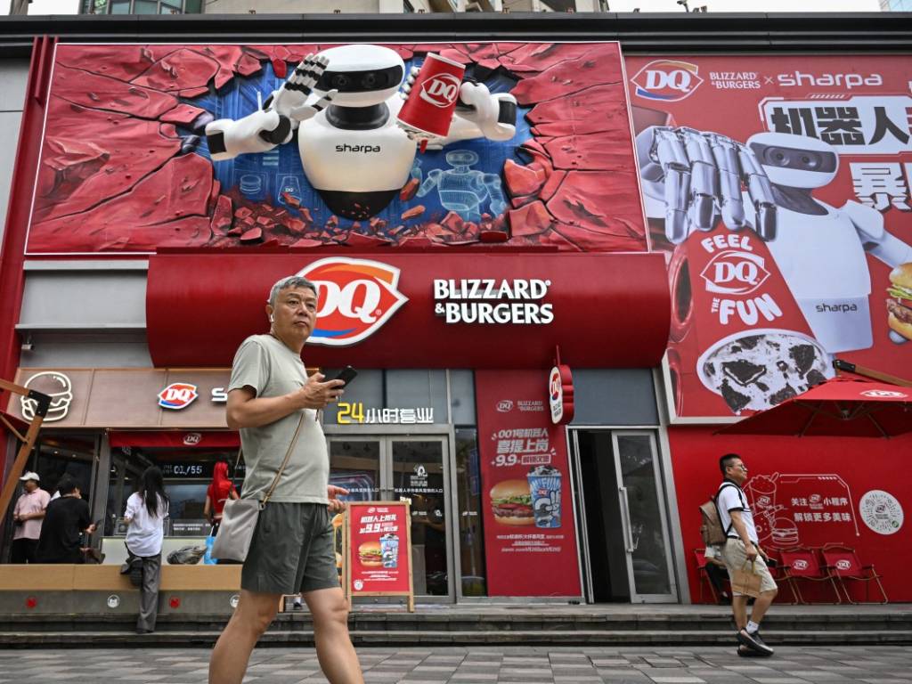 Un hombre caminando por la acera frente al gran ventanal de la heladería Dairy Queen, donde se observa al robot humanoide trabajando adentro.