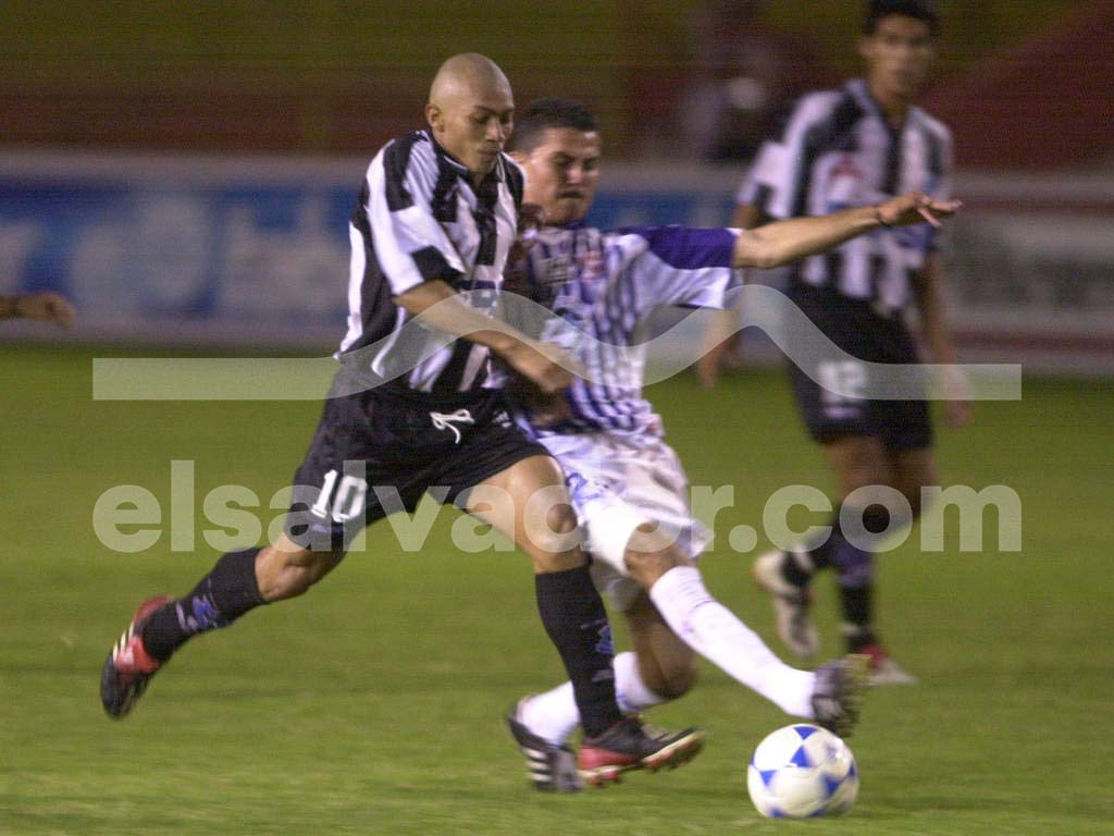 Una fotografía a color que muestra a un jugador de San Salvador FC con su uniforme blanco y azul, disputando un balón con un rival en un estadio con el cielo azul de fondo.