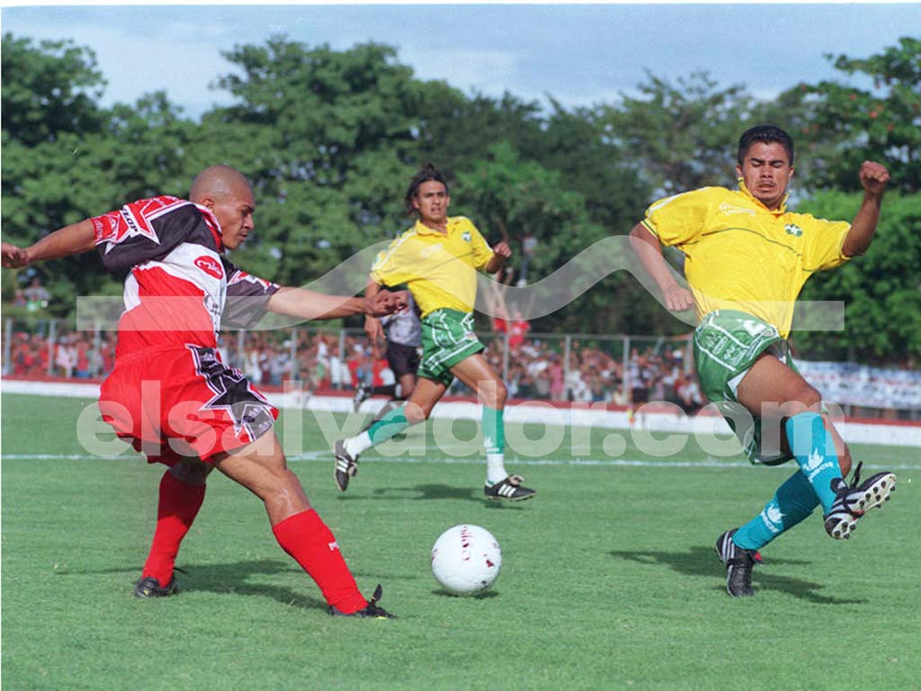 Una foto a color que muestra a un jugador de Santa Clara con su uniforme verde y blanco, disputando un balón con un rival en un campo de juego con vallas publicitarias al fondo.