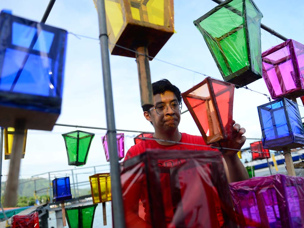 Un hombre joven con gafas y camiseta roja coloca y ajusta faroles de colores (amarillo, verde, azul, magenta) colgados de cables, preparándolos para la procesión nocturna. El fondo muestra un cielo claro y una valla.