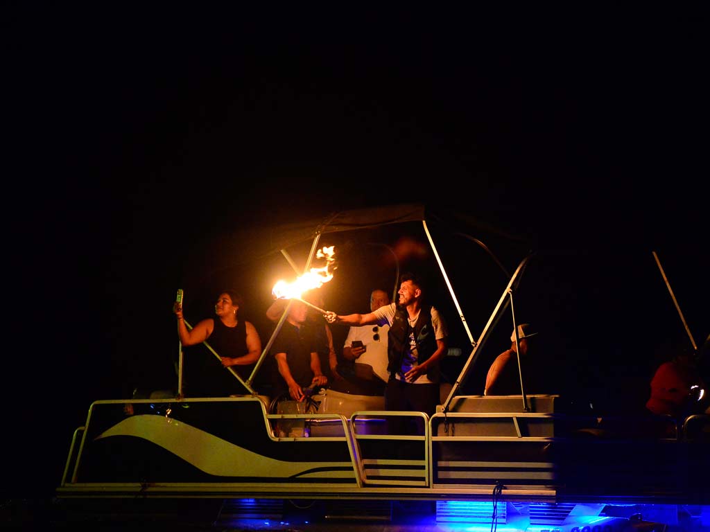 Un grupo de personas en una lancha por la noche. Un hombre sostiene una antorcha encendida, iluminando a los demás a bordo. Se ven luces de colores en la estructura de la lancha y agua oscura alrededor.